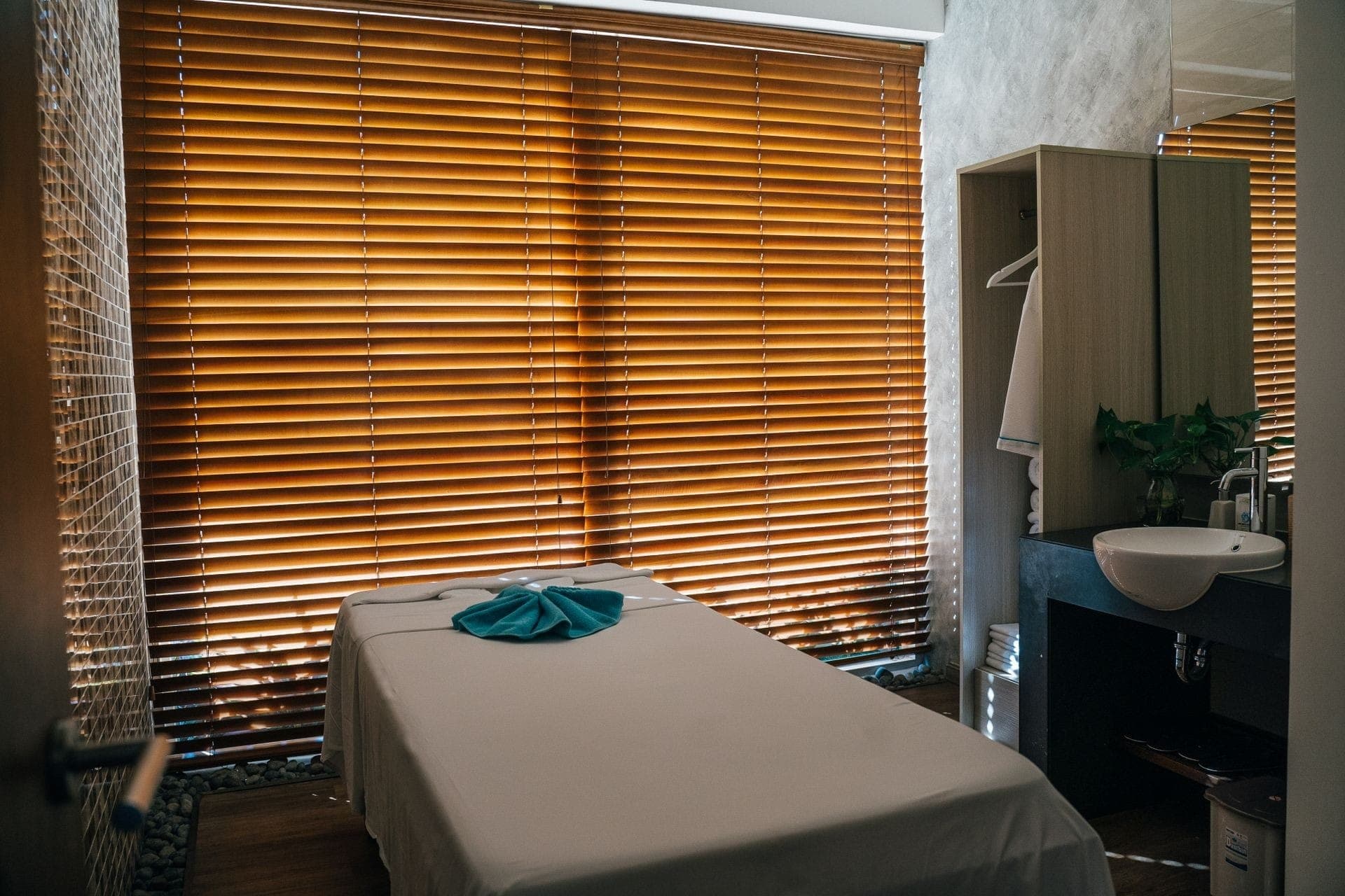 A treatment room with wooden blinds, a made-up massage table and a folded teal towel in warm light